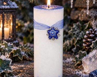 May include: A white cylindrical pillar candle with a lit wick, topped with blue glitter. A sheer blue ribbon is wrapped around the center, secured with a silver-toned star-shaped charm featuring a blue enamel inlay and a central snowflake design.