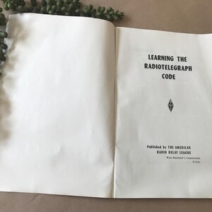 May include: Open book with the title "Learning the Radiotelegraph Code" in black text. Published by The American Radio Relay League, West Hartford, Connecticut, U.S.A. A sprig of green leaves is in the upper left corner.