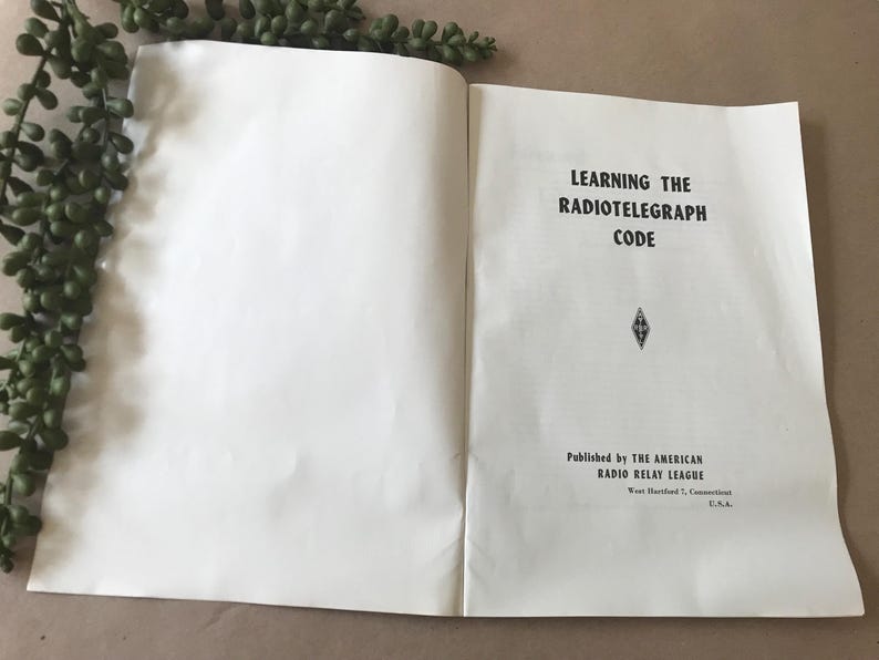 May include: Open book with the title "Learning the Radiotelegraph Code" in black text. Published by The American Radio Relay League, West Hartford, Connecticut, U.S.A. A sprig of green leaves is in the upper left corner.