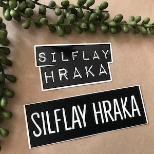 May include: Two black and white rectangular stickers with the text "SILFLAY HRAKA" in a bold, sans-serif font. One sticker is larger than the other. Green plant sprigs are in the background.