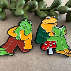 Könnte beinhalten: Zwei Emaille-Anstecknadeln mit Cartoon-Fröschen. Ein Frosch stützt den Kopf auf seine Hand, der andere liest ein Buch, während er auf einem Pilz sitzt. Die Anstecknadeln sind grün, gelb, orange, rot und weiß mit schwarzen Umrissen.