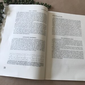May include: Open book with text on pages 22 and 23. The pages are white with black text. The text includes headings like "Automatic Equipment" and "Radiotelegraph Code." A green plant is in the upper left corner.