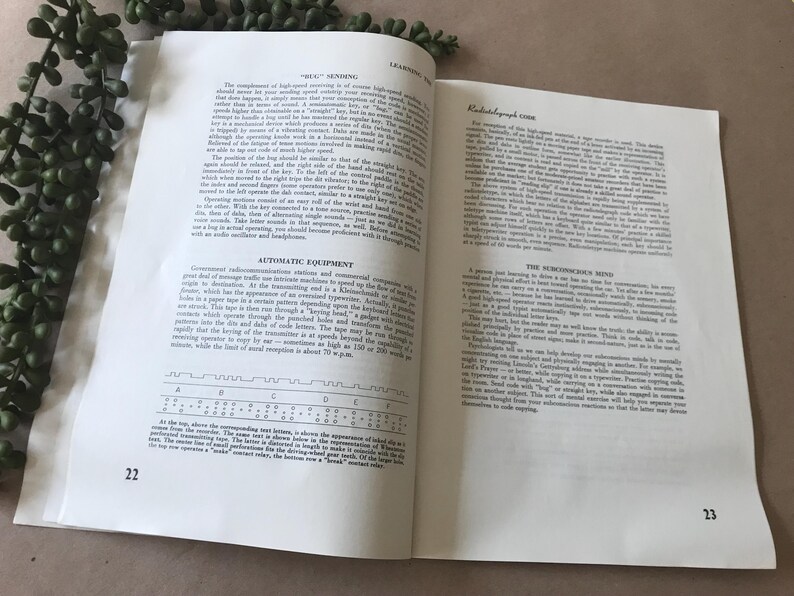 May include: Open book with text on pages 22 and 23. The pages are white with black text. The text includes headings like "Automatic Equipment" and "Radiotelegraph Code." A green plant is in the upper left corner.