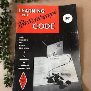 Könnte beinhalten: Vintage-Buch mit dem Titel "Learning the Radiotelegraph Code" mit rotem und schwarzem Einband. Das Buch enthält Text über den Funkbetrieb und den Unterricht im Klassenzimmer. Auf dem Einband ist ein Preis von 50¢ zu sehen.