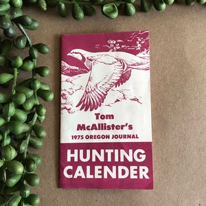 Puede incluir: Un folleto color burdeos con una ilustración blanca de un pájaro en vuelo. El texto de la portada dice "Tom McAllister's 1975 Oregon Journal Hunting Calender".