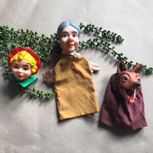 May include: A vintage puppet set comprising three characters: a girl in a red hat, an elderly woman, and a wolf. The puppets have painted faces and fabric clothing. The wolf puppet has a red mouth and tongue.
