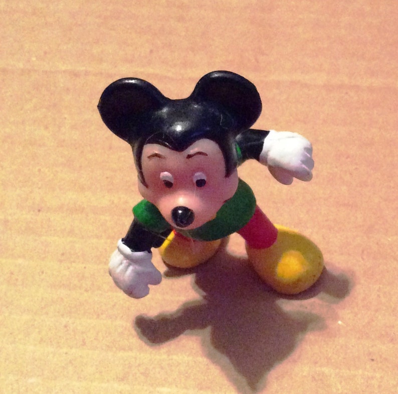 May include: A black and green plastic figurine of Mickey Mouse wearing red shorts and yellow shoes. He is standing with his left arm raised and his right arm bent at the elbow.