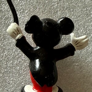 Pu&ograve; includere: Una piccola statuina di Topolino in tenuta da direttore d'orchestra. La figura &egrave; nera con guanti bianchi, la parte inferiore del corpo rossa e una base bianca. Mickey tiene una bacchetta e ha le braccia alzate.