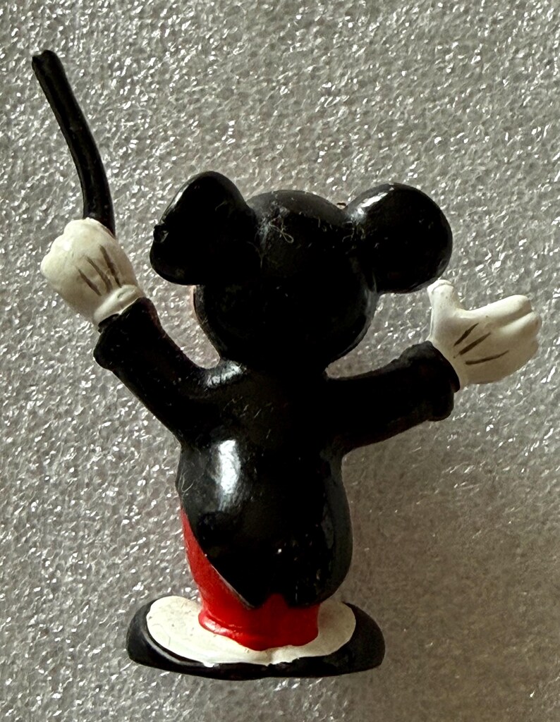 Pu&ograve; includere: Una piccola statuina di Topolino in tenuta da direttore d'orchestra. La figura &egrave; nera con guanti bianchi, la parte inferiore del corpo rossa e una base bianca. Mickey tiene una bacchetta e ha le braccia alzate.