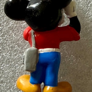K&ouml;nnte beinhalten: Eine Mickey Mouse-Figur von hinten gesehen. Die Figur tr&auml;gt ein rotes Oberteil, eine blaue Hose und gelbe Schuhe. Eine graue Tasche h&auml;ngt &uuml;ber der Schulter. Mickys schwarze Ohren sind sichtbar. Er h&auml;lt die Hand an den Kopf.