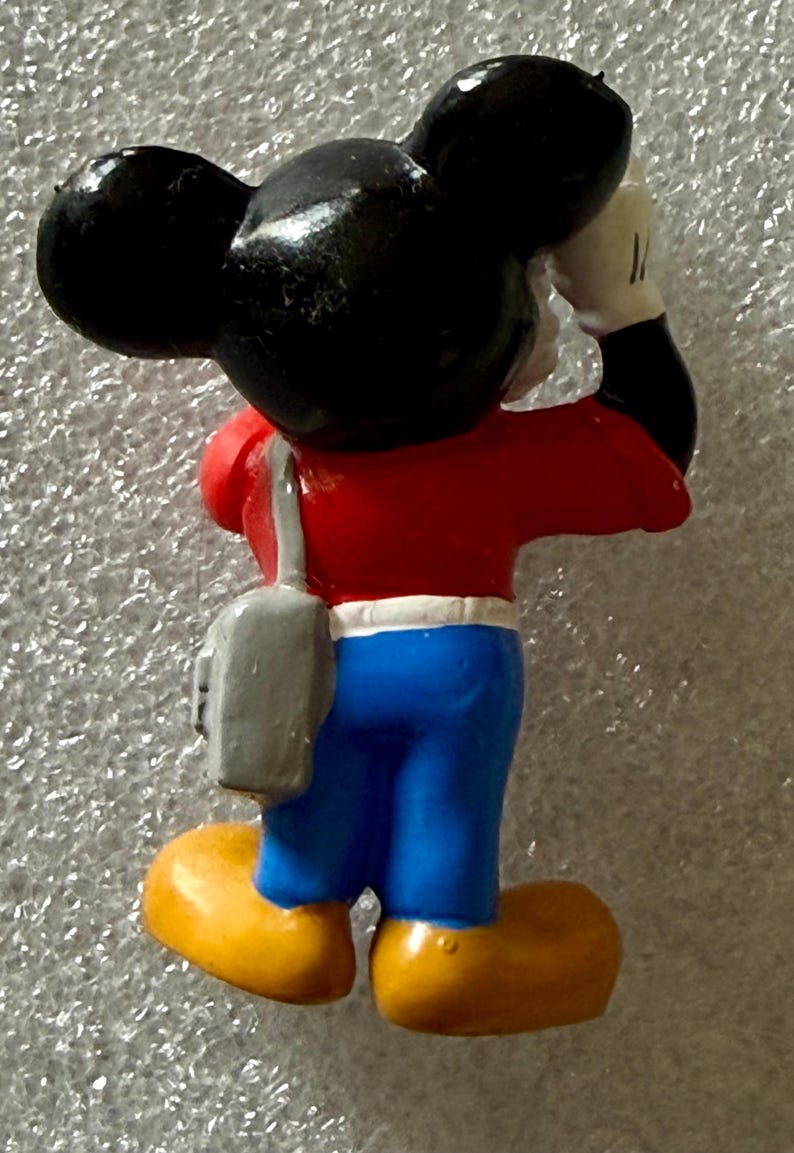 K&ouml;nnte beinhalten: Eine Mickey Mouse-Figur von hinten gesehen. Die Figur tr&auml;gt ein rotes Oberteil, eine blaue Hose und gelbe Schuhe. Eine graue Tasche h&auml;ngt &uuml;ber der Schulter. Mickys schwarze Ohren sind sichtbar. Er h&auml;lt die Hand an den Kopf.