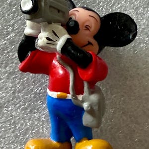 K&ouml;nnte beinhalten: Eine Figur von Micky Maus, die eine graue Videokamera h&auml;lt. Micky tr&auml;gt ein rotes Hemd, blaue Hosen, gelbe Schuhe und wei&szlig;e Handschuhe. Die Figur hat schwarze Ohren und einen l&auml;chelnden Gesichtsausdruck.