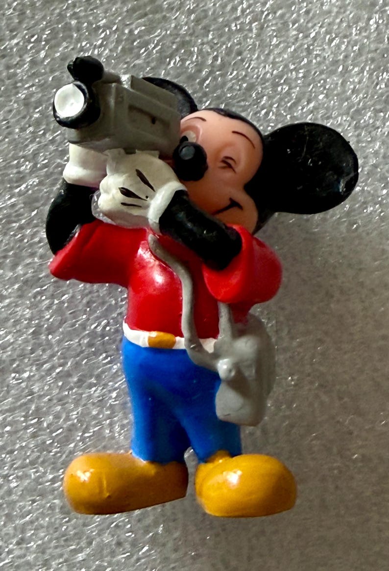 K&ouml;nnte beinhalten: Eine Figur von Micky Maus, die eine graue Videokamera h&auml;lt. Micky tr&auml;gt ein rotes Hemd, blaue Hosen, gelbe Schuhe und wei&szlig;e Handschuhe. Die Figur hat schwarze Ohren und einen l&auml;chelnden Gesichtsausdruck.