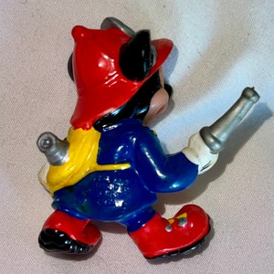 Pode incluir: Uma figurinha de pl&aacute;stico vermelha, azul e amarela do Mickey Mouse vestido de bombeiro. Ele est&aacute; usando um capacete vermelho, uma jaqueta azul e cal&ccedil;as amarelas. Ele est&aacute; segurando uma mangueira de prata na m&atilde;o direita.
