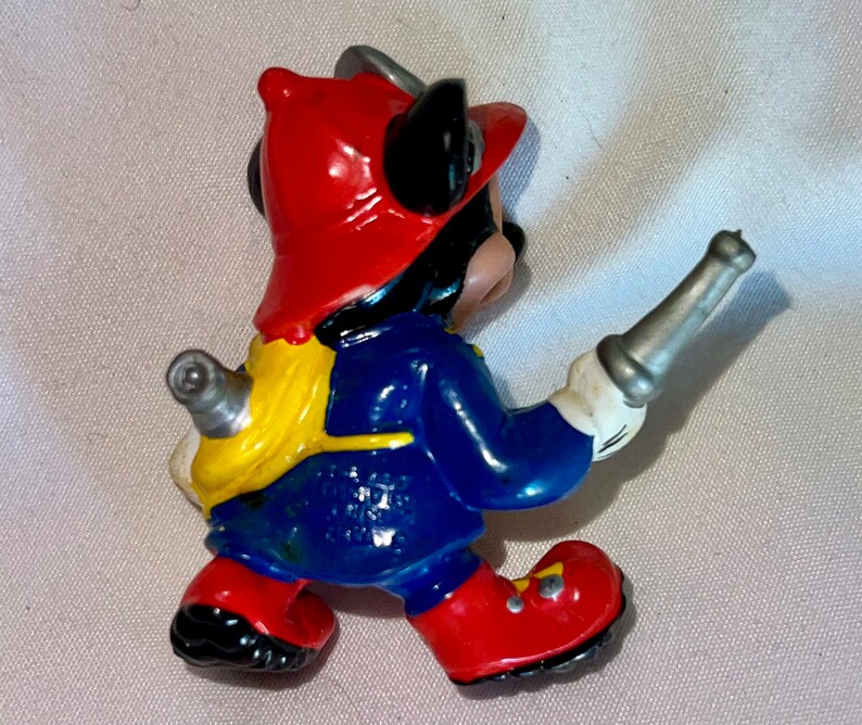 Pode incluir: Uma figurinha de pl&aacute;stico vermelha, azul e amarela do Mickey Mouse vestido de bombeiro. Ele est&aacute; usando um capacete vermelho, uma jaqueta azul e cal&ccedil;as amarelas. Ele est&aacute; segurando uma mangueira de prata na m&atilde;o direita.