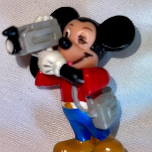 May include: A red and blue Mickey Mouse figurine holding a grey video camera. The figurine is wearing a red shirt, blue jeans, and brown shoes.
