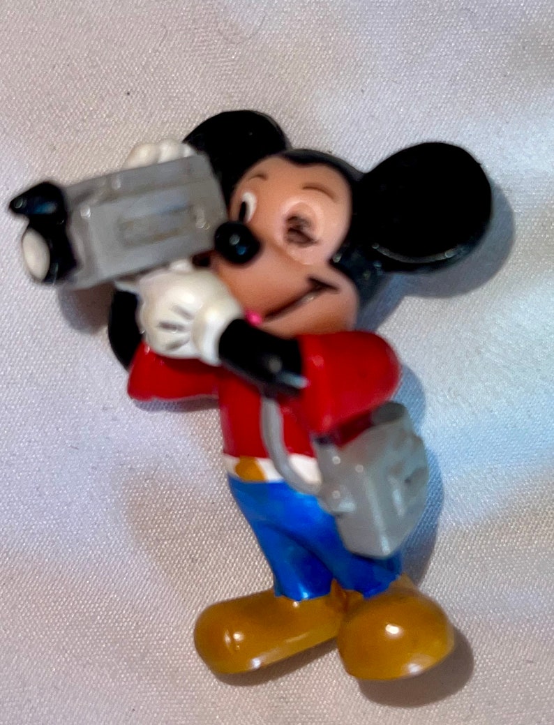 May include: A red and blue Mickey Mouse figurine holding a grey video camera. The figurine is wearing a red shirt, blue jeans, and brown shoes.