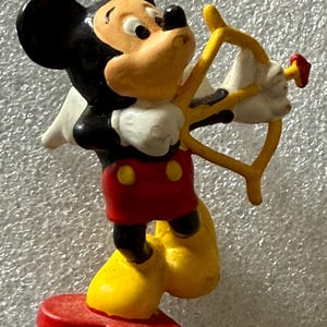 May include: A figurine of Mickey Mouse dressed as Cupid, holding a yellow bow and arrow. Mickey is wearing his signature red shorts with yellow buttons, yellow shoes, white gloves, and a black head with large ears.