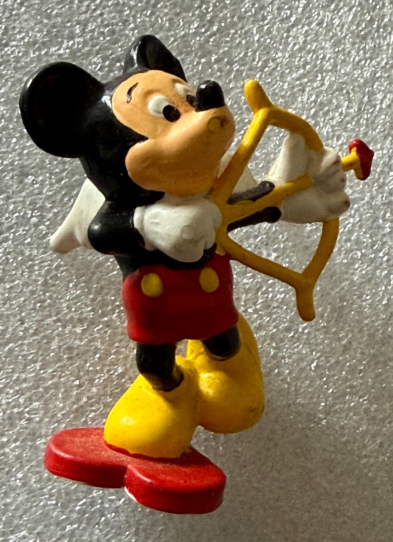 May include: A figurine of Mickey Mouse dressed as Cupid, holding a yellow bow and arrow. Mickey is wearing his signature red shorts with yellow buttons, yellow shoes, white gloves, and a black head with large ears.