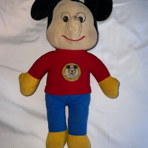 May include: A vintage plush Mickey Mouse doll wearing a red shirt with a yellow Mickey Mouse logo and blue pants. The doll has a large, exaggerated nose and a friendly smile.