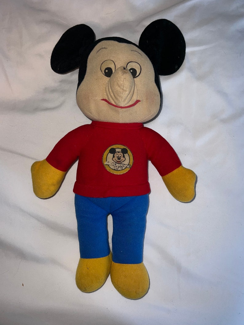 May include: A vintage plush Mickey Mouse doll wearing a red shirt with a yellow Mickey Mouse logo and blue pants. The doll has a large, exaggerated nose and a friendly smile.