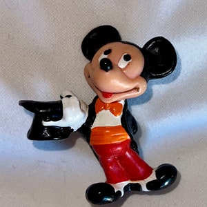 May include: A vintage Mickey Mouse figurine in a magician's outfit. The figure is wearing a black tuxedo, red pants, and a black top hat. Mickey's white gloved hand holds the hat, and he has an orange bow tie.