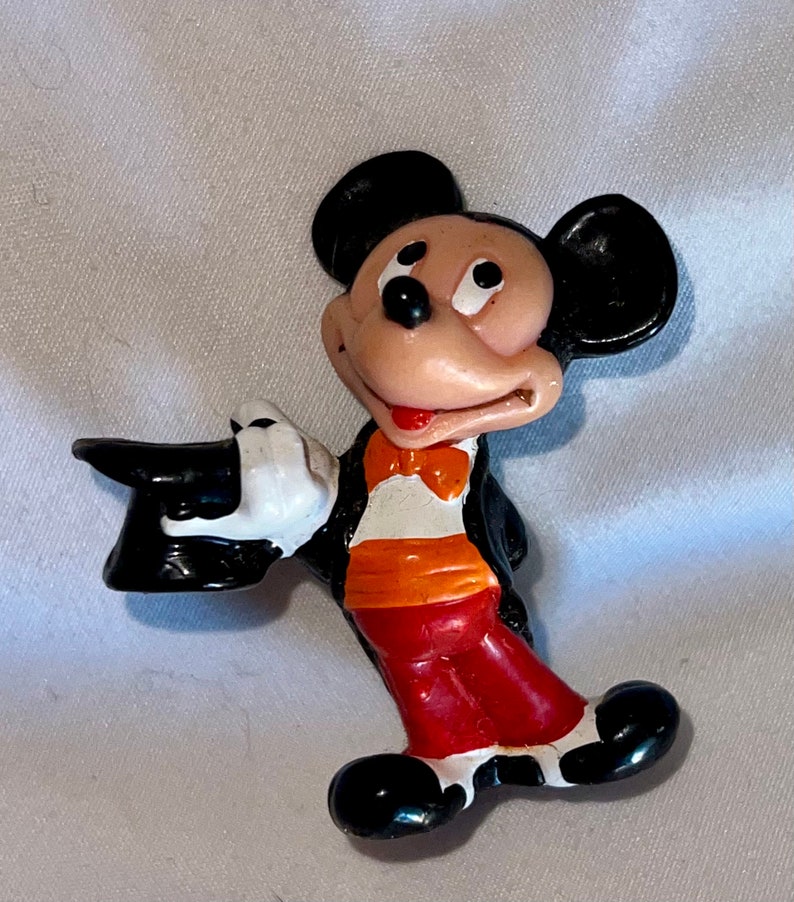 May include: A vintage Mickey Mouse figurine in a magician's outfit. The figure is wearing a black tuxedo, red pants, and a black top hat. Mickey's white gloved hand holds the hat, and he has an orange bow tie.