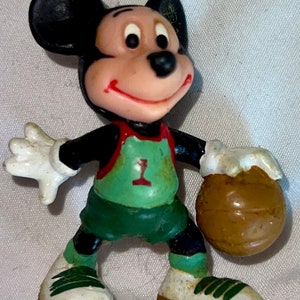 May include: A plastic figurine of Mickey Mouse wearing a green and red basketball uniform with the number '1' on the front. He is holding a brown basketball in his right hand.