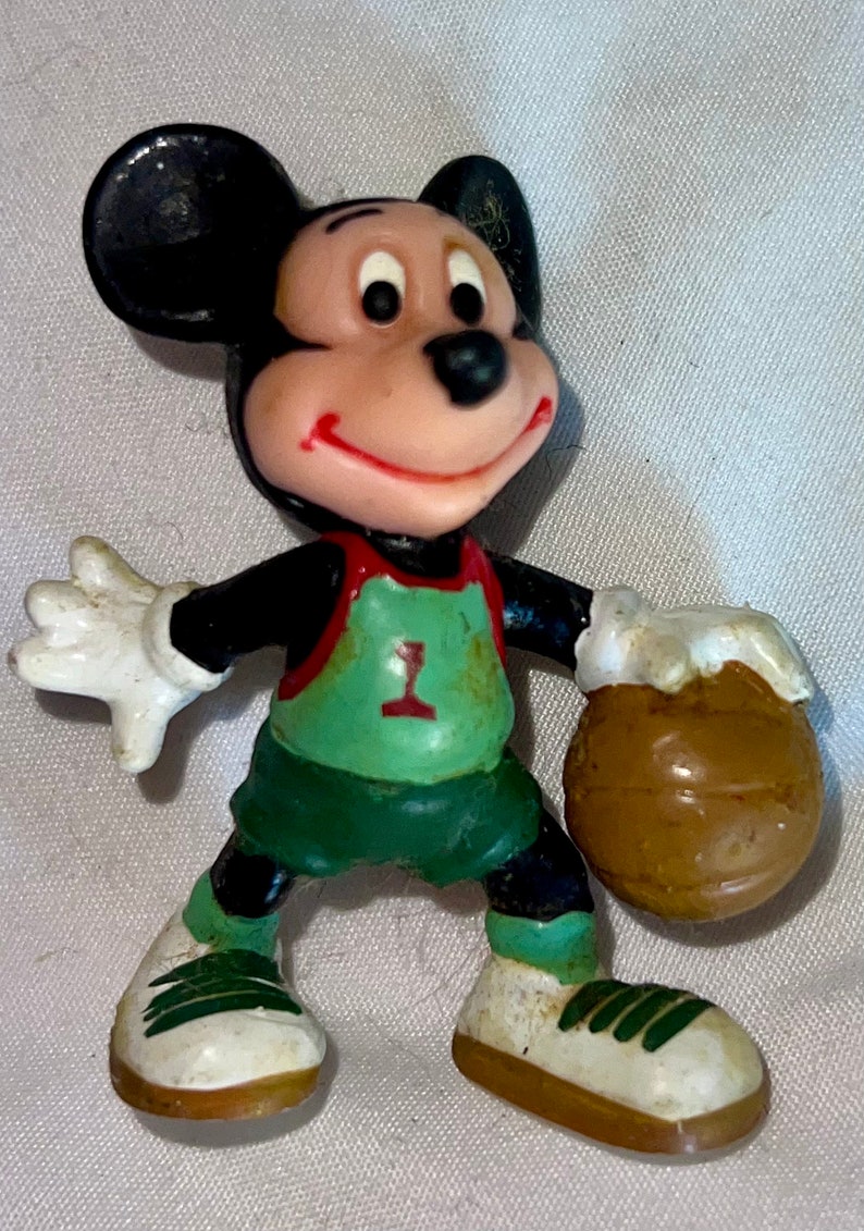 May include: A plastic figurine of Mickey Mouse wearing a green and red basketball uniform with the number '1' on the front. He is holding a brown basketball in his right hand.