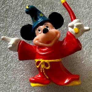 Pu&ograve; includere: Una statuina di Topolino vestito da mago. Mickey indossa una veste rossa con una cintura gialla e un cappello da mago blu con stelle dorate. Tiene in mano una bacchetta rossa e gialla, con un braccio che indica.
