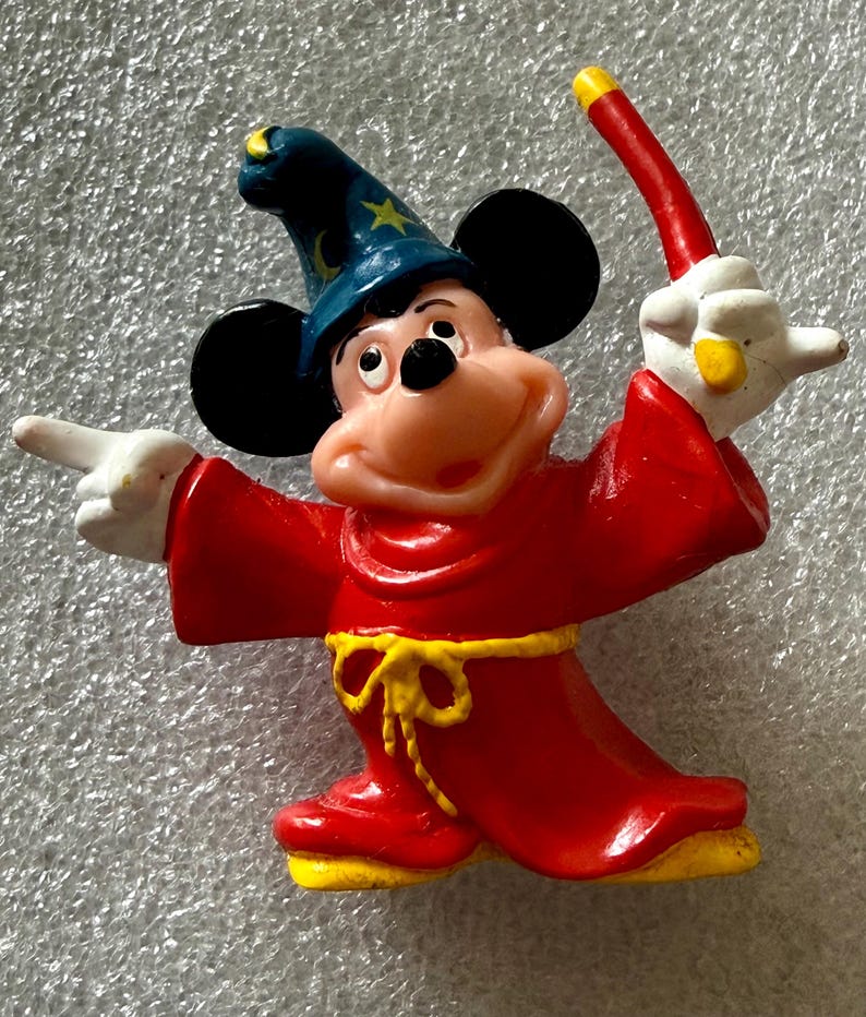 Pu&ograve; includere: Una statuina di Topolino vestito da mago. Mickey indossa una veste rossa con una cintura gialla e un cappello da mago blu con stelle dorate. Tiene in mano una bacchetta rossa e gialla, con un braccio che indica.