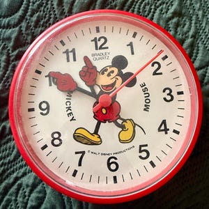 May include: A round Bradley Quartz analog wall clock with a bright red plastic frame and a white face. The clock features black numerals and a central illustration of Mickey Mouse, whose arms serve as the hour and minute hands. Text includes Mickey Mouse, Bradley Quartz, and Walt Disney Productions.