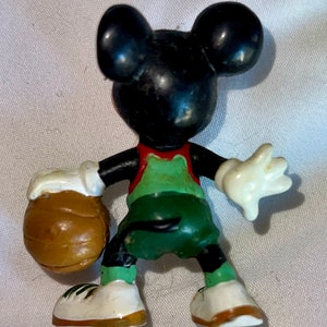 May include: A black and white Mickey Mouse figurine wearing a green and red basketball uniform and holding a brown basketball.