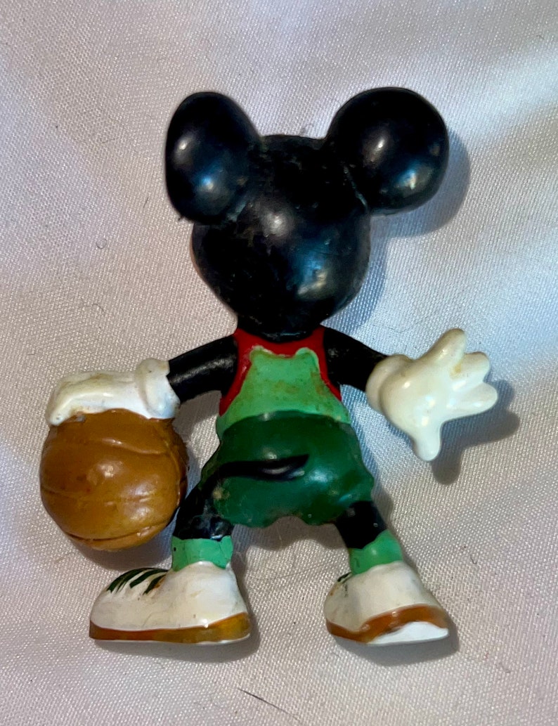 May include: A black and white Mickey Mouse figurine wearing a green and red basketball uniform and holding a brown basketball.