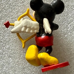 May include: A small figurine of Mickey Mouse as Cupid, viewed from the back. Mickey is black with red shorts, yellow shoes, and a red base. He holds a yellow bow and arrow with a white wing and red heart.
