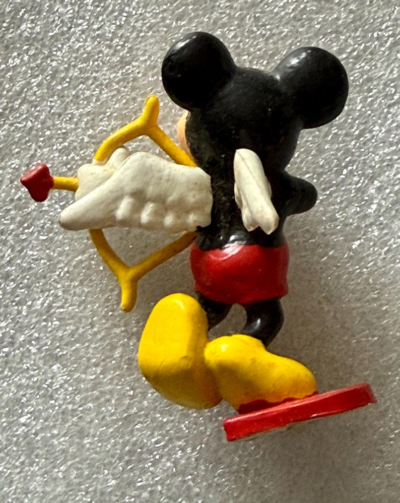 May include: A small figurine of Mickey Mouse as Cupid, viewed from the back. Mickey is black with red shorts, yellow shoes, and a red base. He holds a yellow bow and arrow with a white wing and red heart.