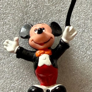 Pu&ograve; includere: Una piccola statuina di Topolino in abito da direttore d'orchestra. Mickey indossa uno smoking nero, pantaloni rossi e un papillon. Tiene in mano una bacchetta nera e ha un'espressione allegra.