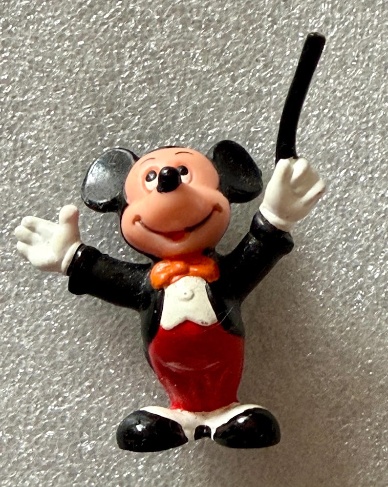 Pu&ograve; includere: Una piccola statuina di Topolino in abito da direttore d'orchestra. Mickey indossa uno smoking nero, pantaloni rossi e un papillon. Tiene in mano una bacchetta nera e ha un'espressione allegra.