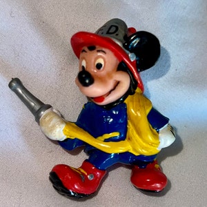Pode incluir: Uma pequena figura de pl&aacute;stico do Mickey Mouse vestido de bombeiro. Ele est&aacute; usando um capacete de bombeiro vermelho e amarelo com as letras "F.D." na frente, uma jaqueta azul, cal&ccedil;as amarelas e botas vermelhas. Ele est&aacute; segurando uma mangueira de inc&ecirc;ndio prateada.