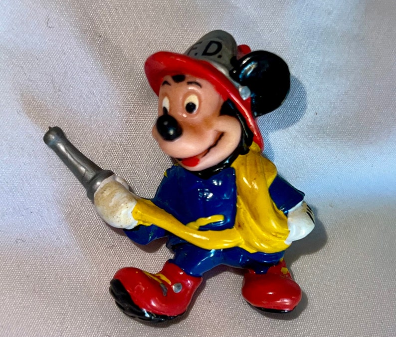 Pode incluir: Uma pequena figura de pl&aacute;stico do Mickey Mouse vestido de bombeiro. Ele est&aacute; usando um capacete de bombeiro vermelho e amarelo com as letras "F.D." na frente, uma jaqueta azul, cal&ccedil;as amarelas e botas vermelhas. Ele est&aacute; segurando uma mangueira de inc&ecirc;ndio prateada.