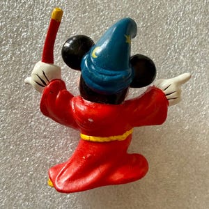 Pu&ograve; includere: Una statuina di Topolino vestito da mago, vista da dietro. La figura indossa una veste rossa con una cintura gialla, un cappello blu con una luna crescente e una stella gialle e guanti bianchi. Mickey tiene in mano una bacchetta rossa e oro.