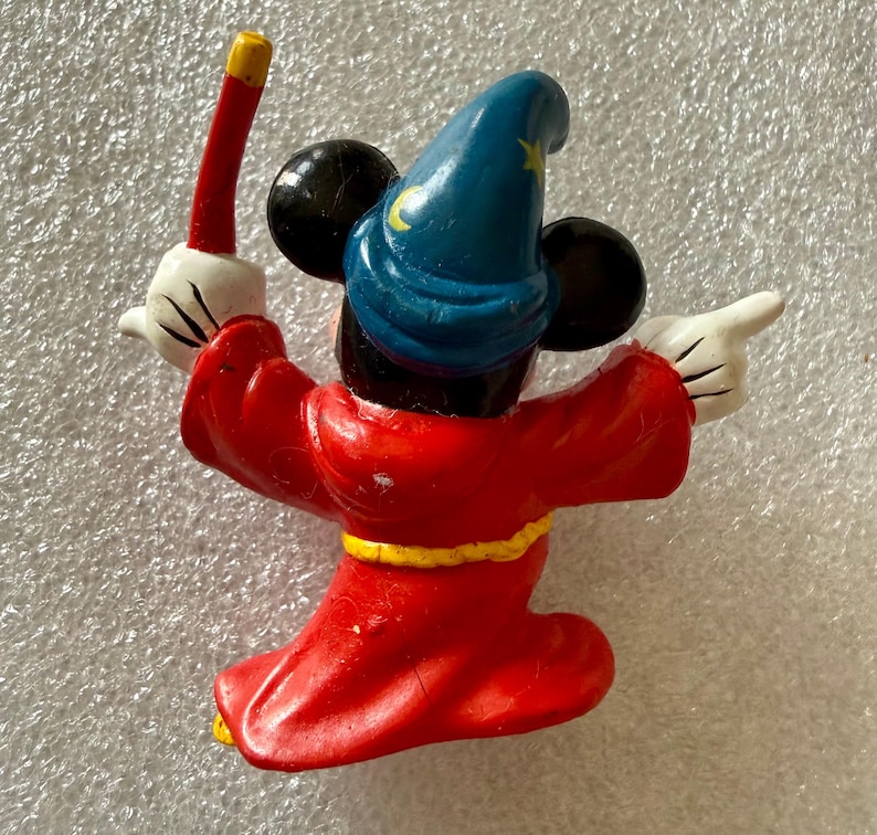 Pu&ograve; includere: Una statuina di Topolino vestito da mago, vista da dietro. La figura indossa una veste rossa con una cintura gialla, un cappello blu con una luna crescente e una stella gialle e guanti bianchi. Mickey tiene in mano una bacchetta rossa e oro.