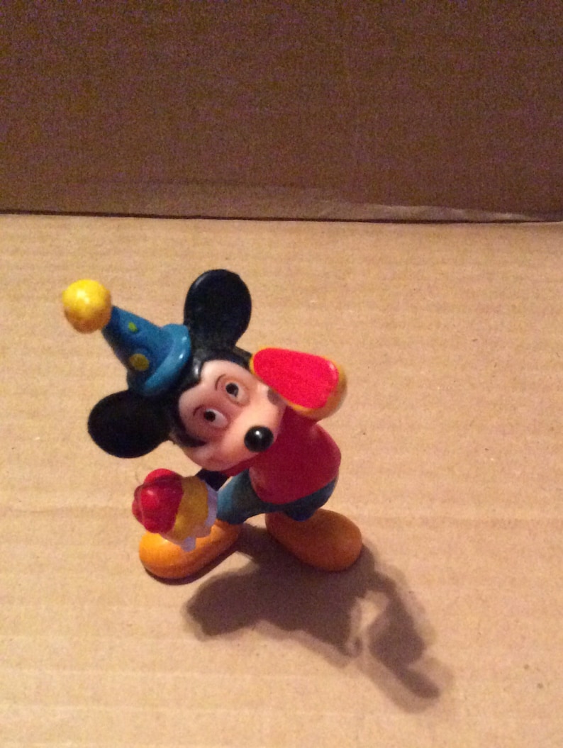 May include: A plastic figurine of Mickey Mouse wearing a blue and yellow hat, a red shirt, and yellow shoes. He is holding a red object in his right hand.
