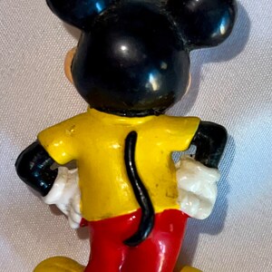 Peut inclure: Une figurine en plastique jaune, rouge et noire de Mickey Mouse. Mickey porte une chemise jaune, un pantalon rouge et des chaussures noires. Il a de grandes oreilles noires et une queue noire.
