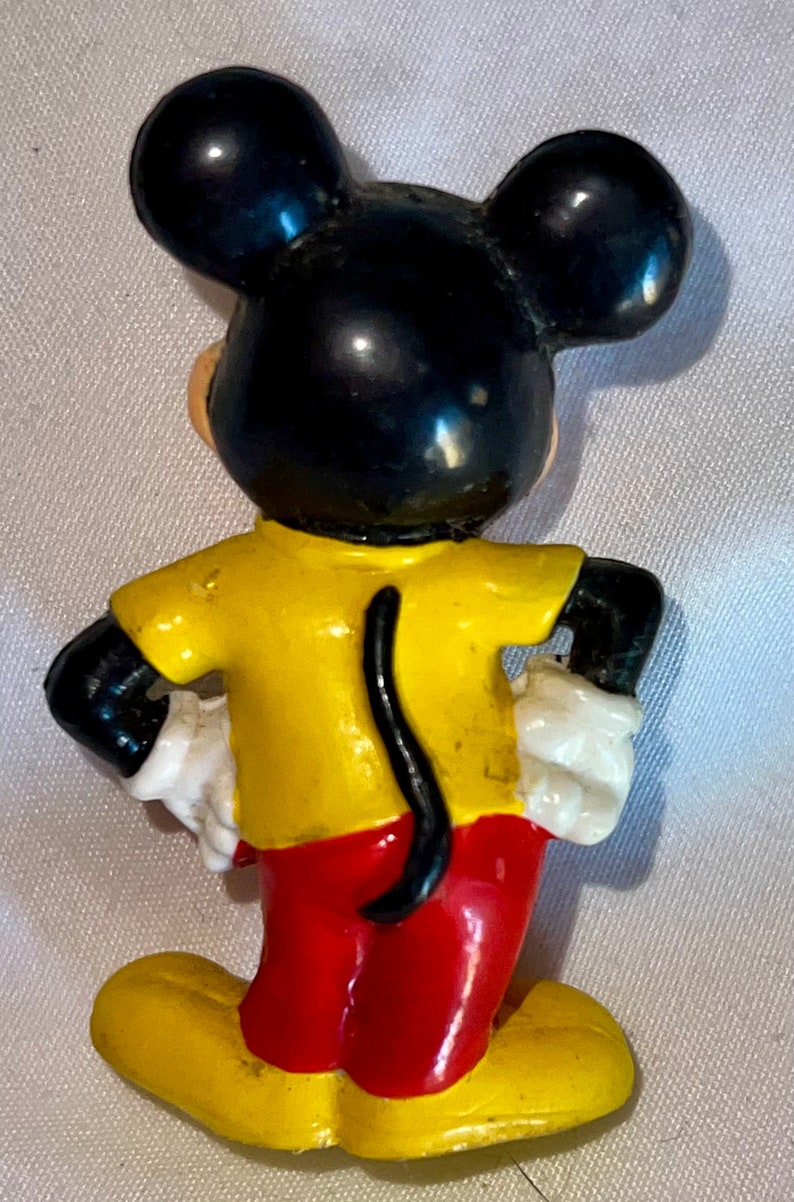 Peut inclure: Une figurine en plastique jaune, rouge et noire de Mickey Mouse. Mickey porte une chemise jaune, un pantalon rouge et des chaussures noires. Il a de grandes oreilles noires et une queue noire.
