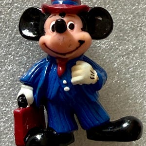 May include: A figurine of Mickey Mouse dressed in a blue suit with pinstripes, a red tie, and a matching hat. He is holding a red briefcase and wearing white gloves and black shoes. The figure has a smiling expression.