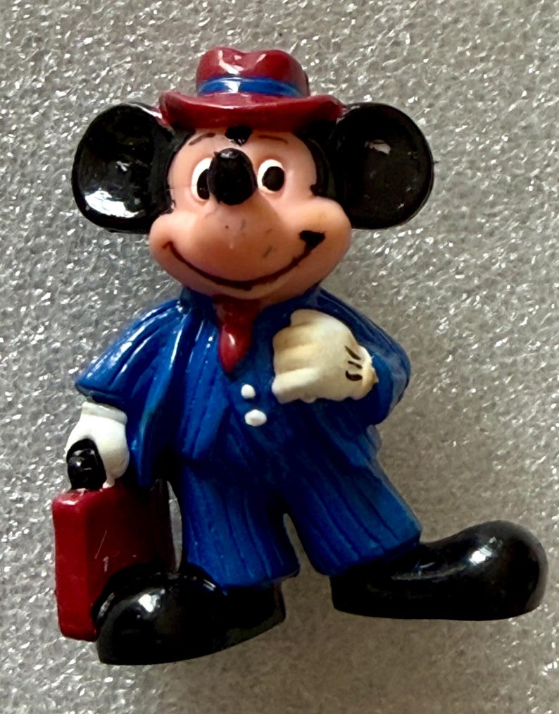 May include: A figurine of Mickey Mouse dressed in a blue suit with pinstripes, a red tie, and a matching hat. He is holding a red briefcase and wearing white gloves and black shoes. The figure has a smiling expression.
