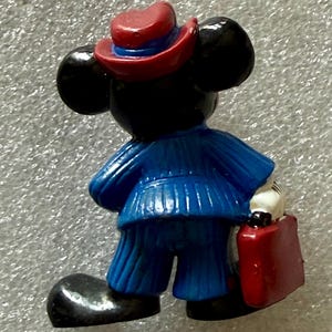 May include: A figurine of a cartoon mouse wearing a blue suit, red hat, and carrying a red briefcase. The mouse has black ears and a white gloved hand. The figurine is standing with its back to the viewer.