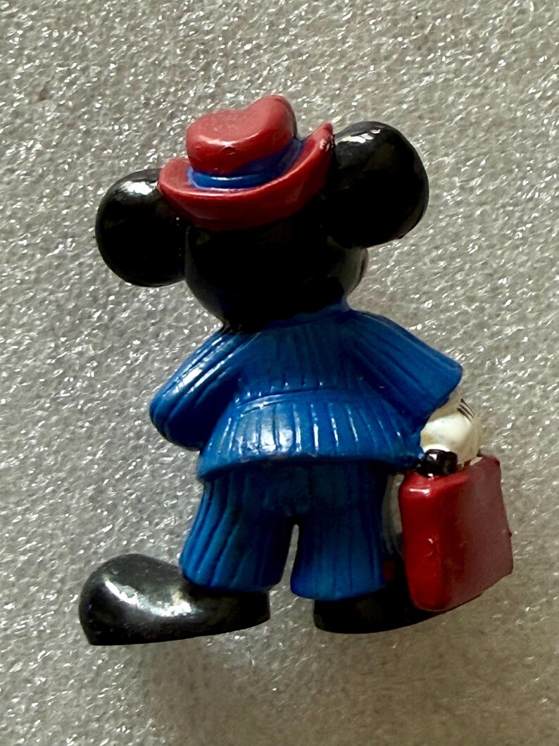May include: A figurine of a cartoon mouse wearing a blue suit, red hat, and carrying a red briefcase. The mouse has black ears and a white gloved hand. The figurine is standing with its back to the viewer.