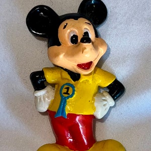 Peut inclure: Une figurine en plastique jaune et rouge de Mickey Mouse portant une chemise jaune avec un ruban bleu sur lequel est inscrit "1".